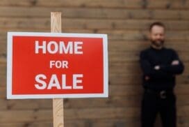 A red Home For Sale sign is in focus in the foreground, while a person with folded arms stands blurred in the background against a wooden wall.