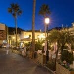 Night view of a lively Spanish plaza with outdoor cafes, palm trees, and historic buildings illuminated by street lamps, creating a charming atmosphere.